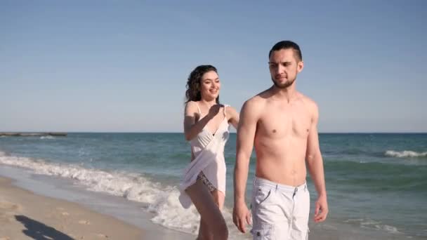 积极的年轻朋友在夏天的休息，女孩和男人一起跑着跳着回来，恋人在海滩上，风吹着女人的头发, — 图库视频影像