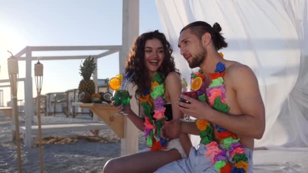 Przyjaciele na plaży, kolorowe wieńce na szyi kochający siedzący na brzegu morze, mężczyzna i dziewczyna na podświetleniu promieni słońca, szczęśliwa para — Wideo stockowe