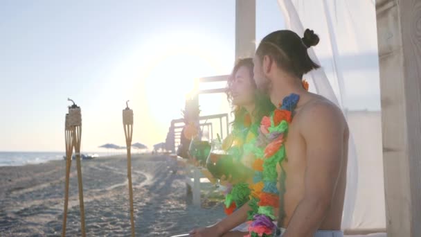 Frau sein Mann zeigt mit der Hand in die Ferne, hawaiianische Lei am Hals junge Leute am Ufer des Ozeans, Gegenlicht der Sonnenstrahlen — Stockvideo