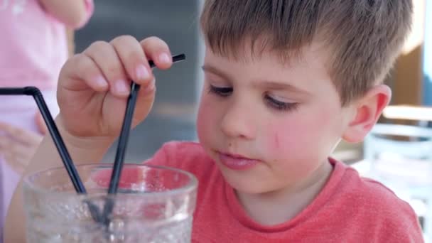 Pojke dricker dryck genom halm från glas sitter vid bordet i mysiga gatan café, Vackra barn gör mellanmål i restaurang, rolig unge äter — Stockvideo