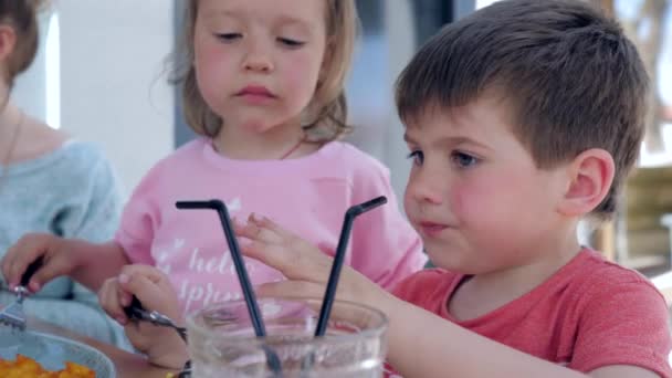Дети пьют сок через солому, вместе из одного стакана, сидя за столом в уютном уличном кафе, красивый брат и сестра делают закуски — стоковое видео
