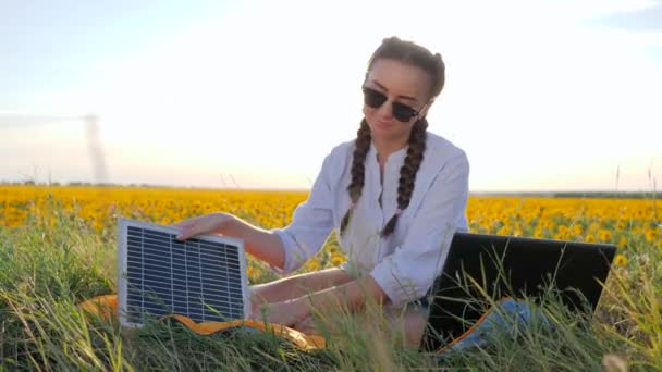 Chica recarga el ordenador portátil utilizando la batería solar en el campo de los girasoles, mujer joven aplicando paneles solares fotovoltaicos al aire libre — Vídeos de Stock