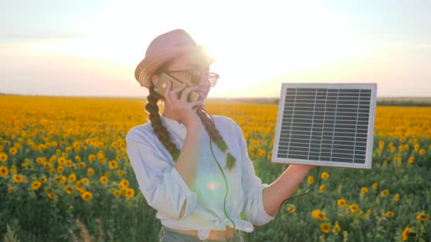 Menina fala por telefone celular e detém painel solar no campo de fundo de girassóis, jovem com telefone e bateria solar — Vídeo de Stock