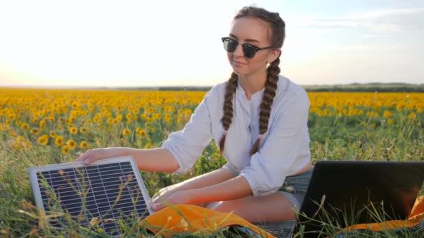 Nová technologie, dívka dobíjí notebook pomocí solární baterie na poli slunečnic, mladá žena nanáší solární fotovoltaické panely — Stock video