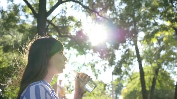 Söt flicka håller glasflaska i handen och dricker kallt rent vatten i parken på bakgrundsträd i bakgrundsbelysning — Stockvideo