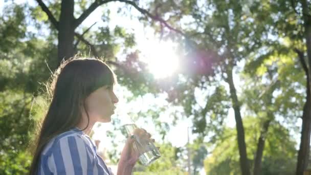 Jeune femme tient bouteille en verre dans sa main et boit de l'eau minérale sur les arbres de fond dans le contre-jour — Video