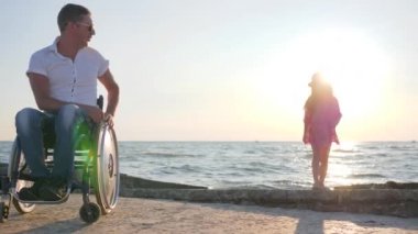 Tekerlekli sandalyedeki engelli adam, arka planda, deniz kenarında zıplayan çocuğu yavaş çekimde alkışlıyor.