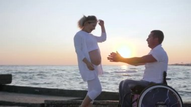 Günbatımında tekerlekli sandalyede sakat kocası olan hamile bir kadın. Mutlu engelli, koca göbekli bir kadını okşuyor.