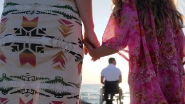 Hamile karısı ve kızı olan engelli bir adam, el ele tutuşan kız ve anne, anne ve kızı tekerlekli sandalyede oturan babaya el ele tutuşuyorlar.