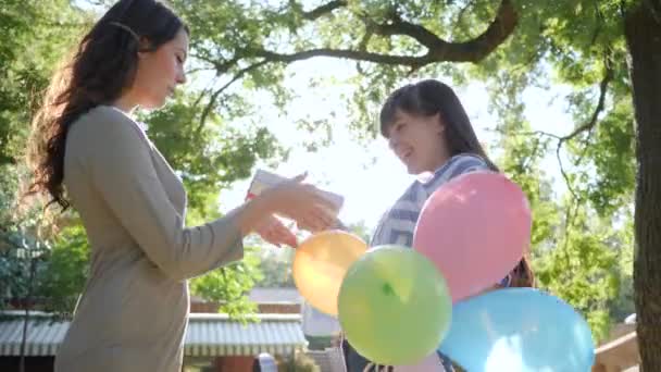 Érzelmek a napsütéses napon, vidám nő színes lufikkal gratulál barátja boldog születésnapot a parkban — Stock videók