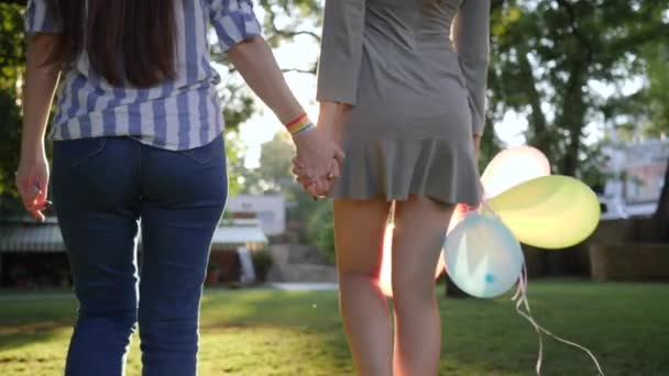 Девушка с надувными воздушными шарами держит за руку женщину с браслетом ЛГБТ на заднем плане зеленые деревья в подсветке — стоковое видео
