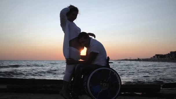 Инвалид слушает брюшную полость жены вечером, инвалид супруга в инвалидной коляске с беременной женщиной на закате — стоковое видео