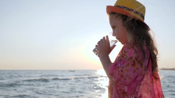 Liten flicka i hatt dricker rent vatten nära havet med vågor i bakgrundsbelysning, närbild söt barn håller kopp i händerna vid solnedgången — Stockvideo
