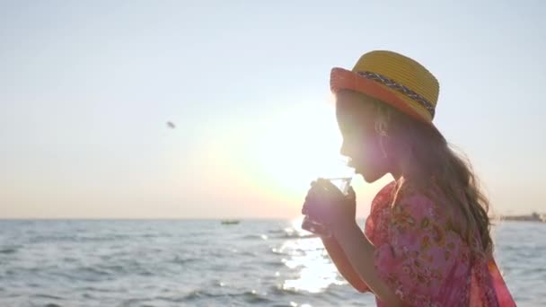 Mãe dá água sua filha ao ar livre, menina bebe água limpa perto do mar, criança bonita no backlight mantém copo — Vídeo de Stock