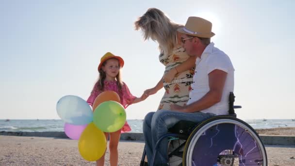 Ebeveynler hamilelikten, tekerlekli sandalyedeki engelli insandan bahseder. Küçük kız, plajda anne ve babasının tekerlekli sandalyesini dinler. — Stok video