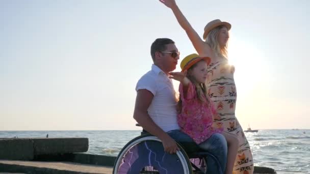 Инвалид летом, семья играет вместе на фоне голубого неба, маленькая девочка сидит на папочке в инвалидной коляске с вытянутыми руками — стоковое видео