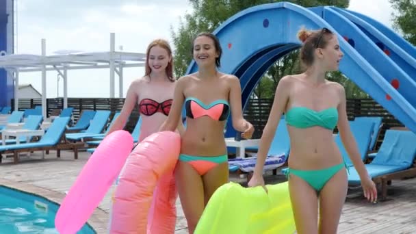 Flickor i bikini går på träpiren bredvid poolen med rosa gummiringar, flickvänner i baddräkt med bad — Stockvideo