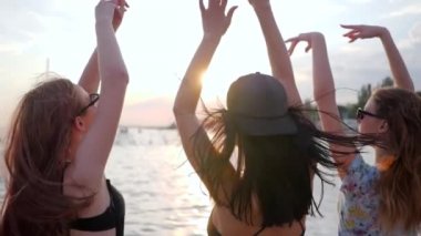 Parti, kumsalda gün batımında dans eden arkadaşlar, kız arkadaşlar deniz kenarında kollarını kaldırıyor, genç kadınlar dans ediyor.