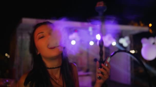 Jovem mulher fumaça narguilé e segurando móvel em festa em luzes, close-up bela fêmea inala sabor do tabaco — Vídeo de Stock