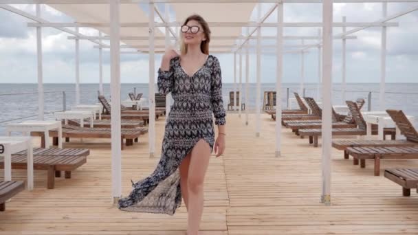 Vrouw in zonnebril loopt langs steiger in de buurt van zee, langharig meisje in de ontwikkeling van kleding aan het water, jonge vrouw — Stockvideo