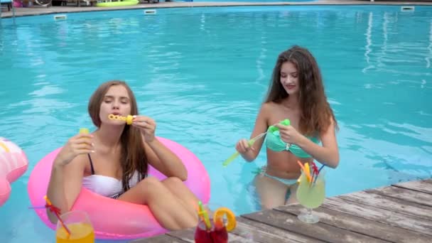 Подружки в купальнике дуют мыльные пузыри в бассейне, милые девушки сидят у надувного кольца в бассейне и делают пузыри , — стоковое видео