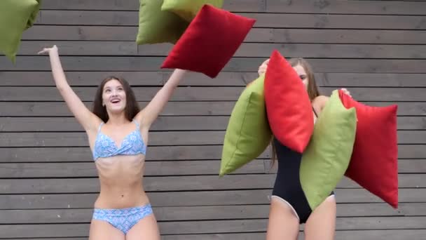 Des amis riants s'amusent sur le mur en bois de fond, des copines en maillots de bain jettent un coussin multicolore — Video