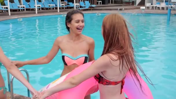 Amigos riendo tomados de la mano bailan en la piscina con agua azul, verano, niñas alegres con anillos inflables rosados — Vídeos de Stock