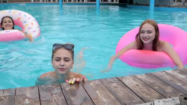 Meninas de retrato nadando com anéis infláveis cor-de-rosa na beira da piscina, amigos nadam na piscina com água azul — Vídeo de Stock