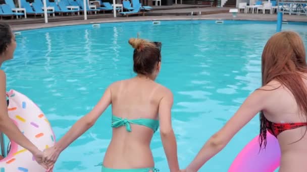 Amici vista posteriore tenendo le mani saltare in piscina con acqua blu, gioiose ragazze con anelli gonfiabili rosa divertirsi — Video Stock