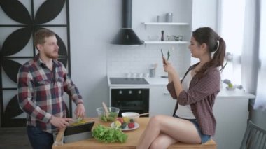 Neşeli kız masada oturuyor ve cep telefonuyla komik adam fotoğrafı çekiyor. Brunch için yararlı yemekler pişiriyor.