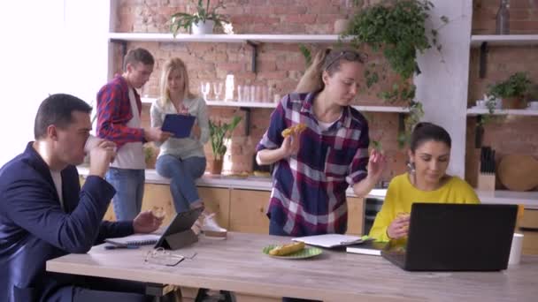 Equipo de trabajo de jóvenes empresarios exitosos están comiendo y trabajando con tabletas y computadoras portátiles en la cocina durante la fabricación de un nuevo proyecto creativo en la oficina — Vídeos de Stock