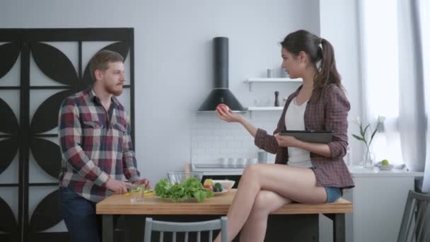 Atrakcyjny brodaty samiec przygotowuje pyszne zdrowe sałatki ze świeżych warzyw i zieleni, a samica siedzi na stole z tabletką w ręku i rozmowy — Wideo stockowe