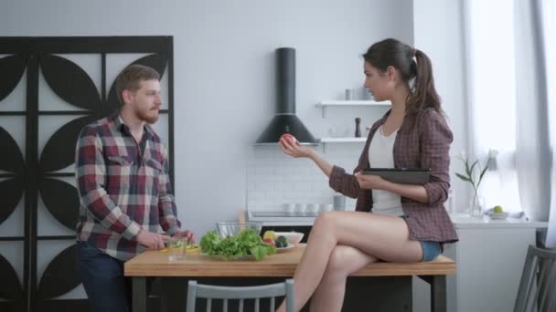 Atractivo barbudo macho prepara deliciosa ensalada saludable de verduras frescas y verduras y hembra se sienta en la mesa con la tableta en la mano y habla — Vídeos de Stock