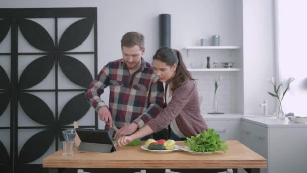 Kontrola kalorii, szczęśliwa dziewczyna z facetem przygotowuje zdrowe jedzenie warzyw i zieleni zgodnie z planem diety dla zdrowia na stole kuchennym przy użyciu nowoczesnej tabletki z przepisem — Wideo stockowe