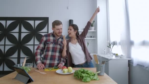 Familia alegre, esposa atractiva con el marido bailando y divirtiéndose mientras cocina comida saludable de verduras en la cena para el bienestar de acuerdo con el plan de dieta en la cocina — Vídeo de stock