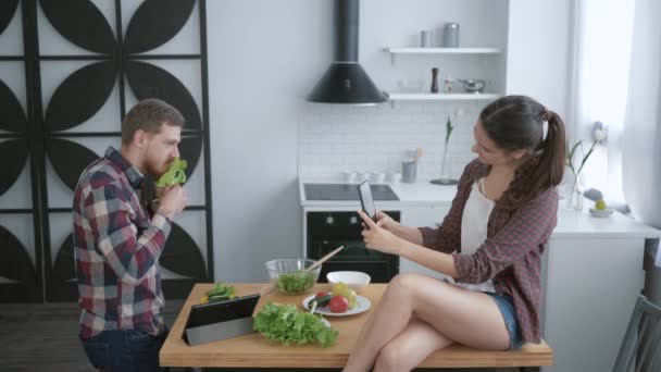 Pazzo ragazzo incredibile scherzare e fare smorfie con verdure e ragazza scatta foto al telefono mentre seduto sul tavolo in cucina mentre cucina sano mangiare — Video Stock