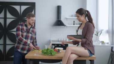 Sağlıklı beslenme, çekici erkek, akşam yemeğinde yararlı vejetaryen yiyecekler hazırlar ve kadın, elinde tabletle mutfak masasında oturur ve yemek pişirme tavsiyesi verir.