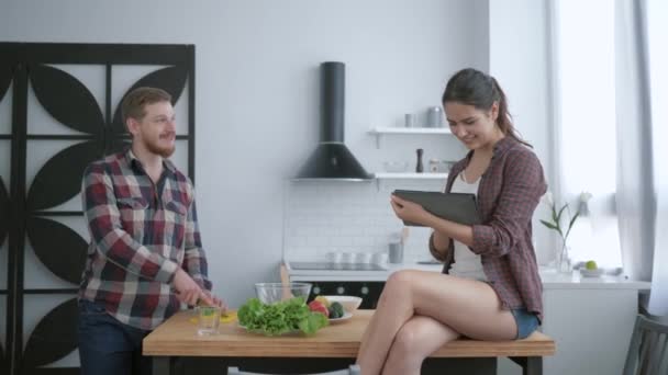 План дієти, щасливий хлопець готує смачний здоровий салат зі свіжих овочів і зелені, а дівчина сидить на столі з таблетками в руках і розмовах — стокове відео