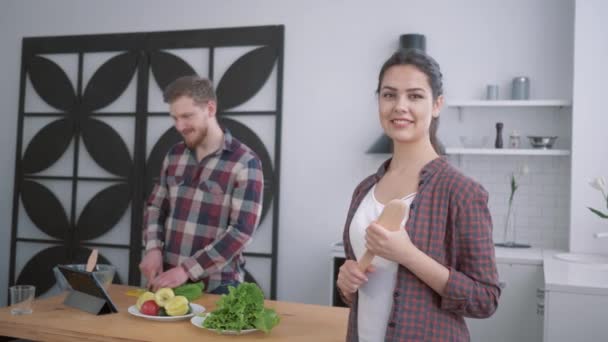 Retrato de esposa feliz en la cocina, mujer satisfecha mira al marido que prepara una alimentación saludable de verduras para el almuerzo útil utilizando tableta en la mesa de cocina — Vídeo de stock