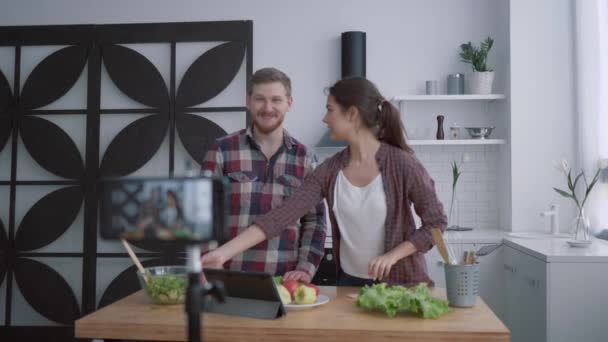 Beslenme blogu, vloggerlar erkek ve kadın mutfakta sebze ve yeşillerle sağlıklı bir kahvaltı hazırlarken, kameralı akıllı telefon sosyal ağlardaki abonelere video kaydediyor. — Stok video