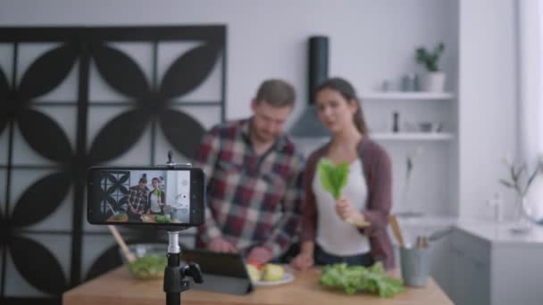 Zayıflatma blogu, vloggerlar erkek ve kadın mutfaktaki sebzeler ve yeşilliklerle sağlıklı bir kahvaltı hazırlarken, kamera cep telefonu sosyal ağlardaki abonelerin videolarını kaydediyor — Stok video