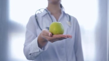 Ellerinde yeşil elma olan doktor, beyaz arka planda sağlıklı beslenme tavsiyesi veren kameraya uzanıyor.