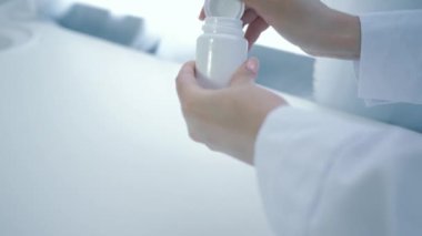 Sağlık hizmetleri, diyetisyen eller vitamin şişesini açar ve sarı yuvarlak hapları beyaz masaya döker.