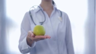 Ellerinde yeşil elma olan profesyonel diyetisyen, beyaz arka planda sağlıklı beslenme tavsiyesi veren kameraya uzanıyor.