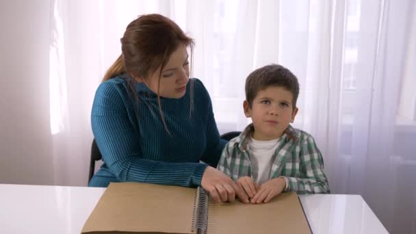 Schwierige Schulbildung für blinde Kinder, junge Lehrerin lehrt sehbehinderten Jungen, Brailleschrift-Bücher mit Schriftzeichen zu lesen, die am Tisch im Zimmer sitzen — Stockvideo