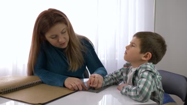 Educación de los niños ciegos, madre enseña niño niño a escribir braille sentado en la mesa en habitación luminosa — Vídeos de Stock