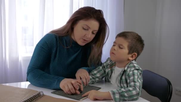 Onderwijs Visueel gehandicapten kinderen, moeder leert blinde zoon jongen om braille karakters lettertype zitten aan tafel in lichte kamer te schrijven — Stockvideo