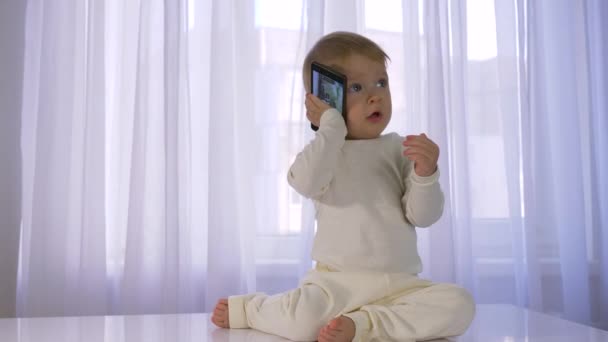 Infância moderna, lindo bebê engraçado é jogado e falando no telefone celular em quarto brilhante — Vídeo de Stock