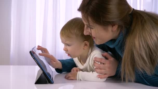 Современные технологии в семейных отношениях, счастливый ребенок с мамой играют с планшетом в светлой комнате — стоковое видео