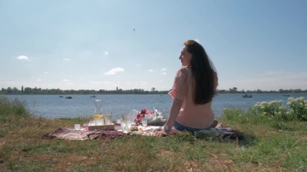 Schöne junge Frau trinkt Wasser aus Glas beim Picknick auf dem grünen Rasen am Fluss und genießt die Ruhe — Stockvideo